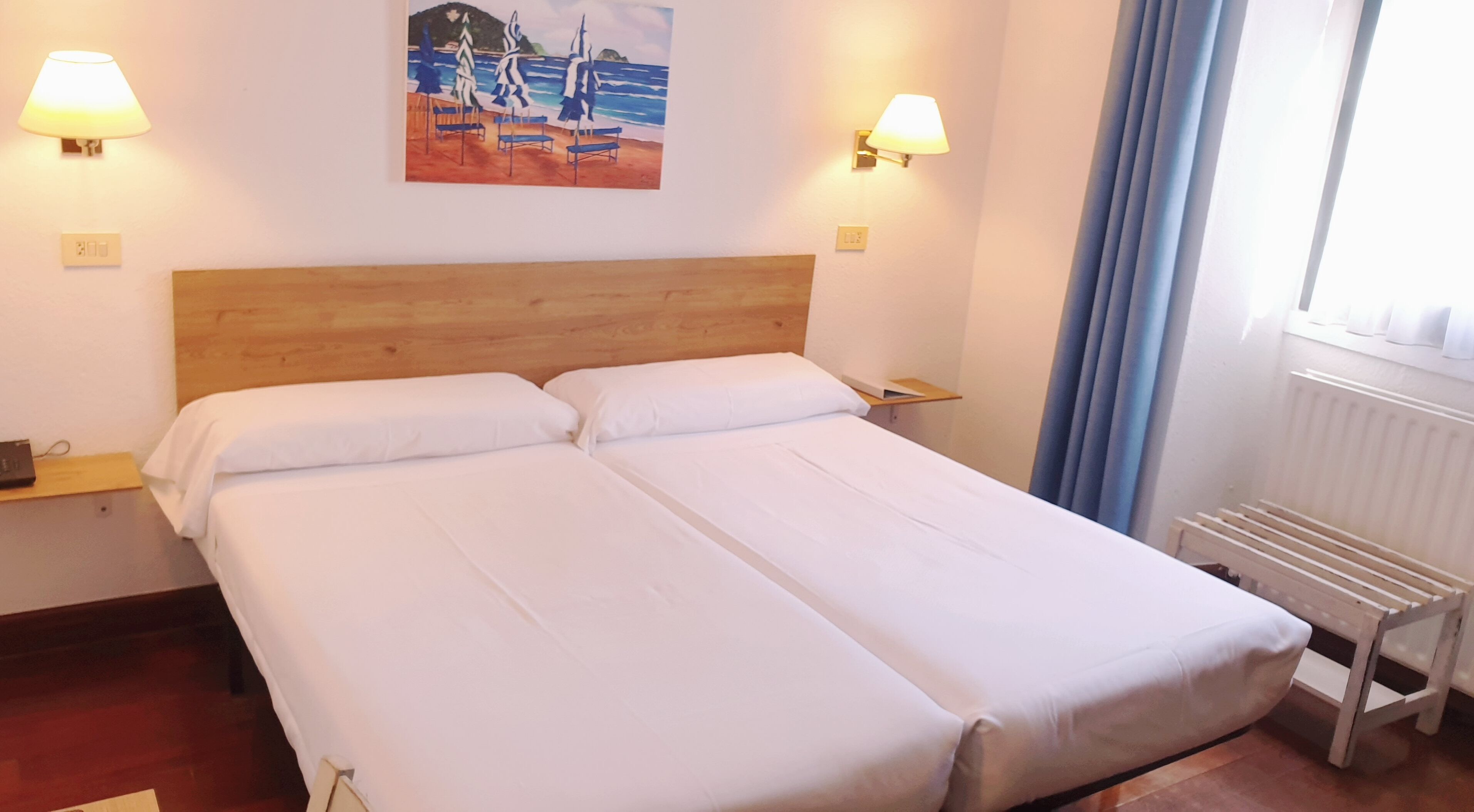 A double bed with white linens, a wooden headboard, two bedside lamps, and a painting of a beach scene on the wall.
