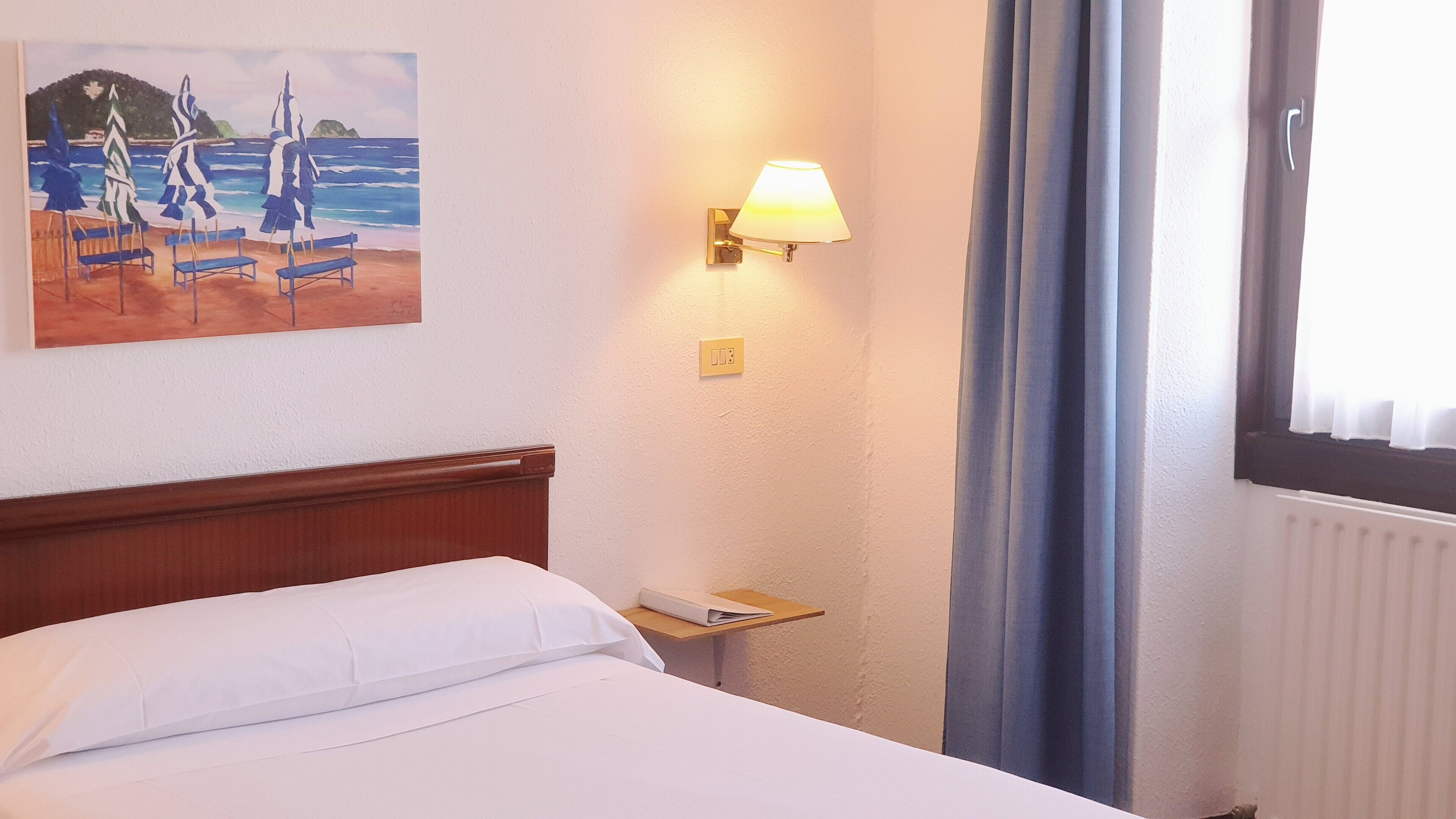 A hotel room with a bed, a nightstand, a lamp, a painting of a beach scene, and a window with curtains.