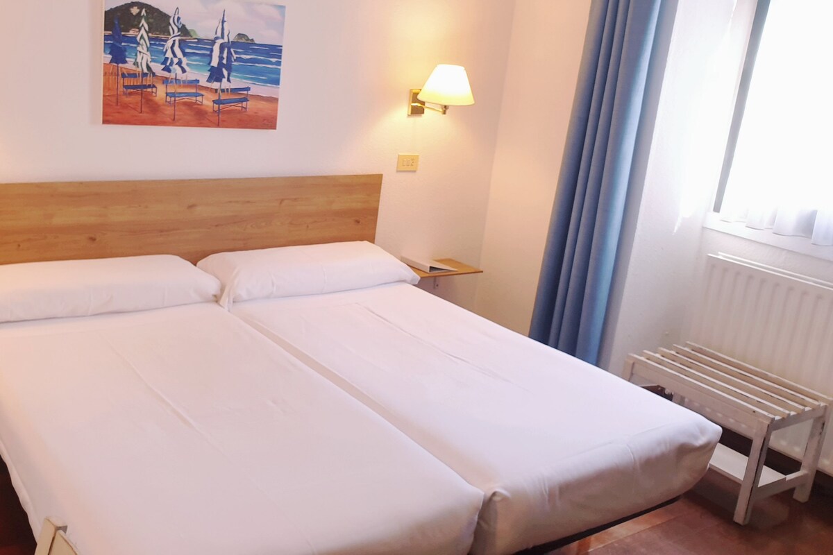 A double bed with white linens, a wooden headboard, two bedside lamps, and a painting of a beach scene on the wall.