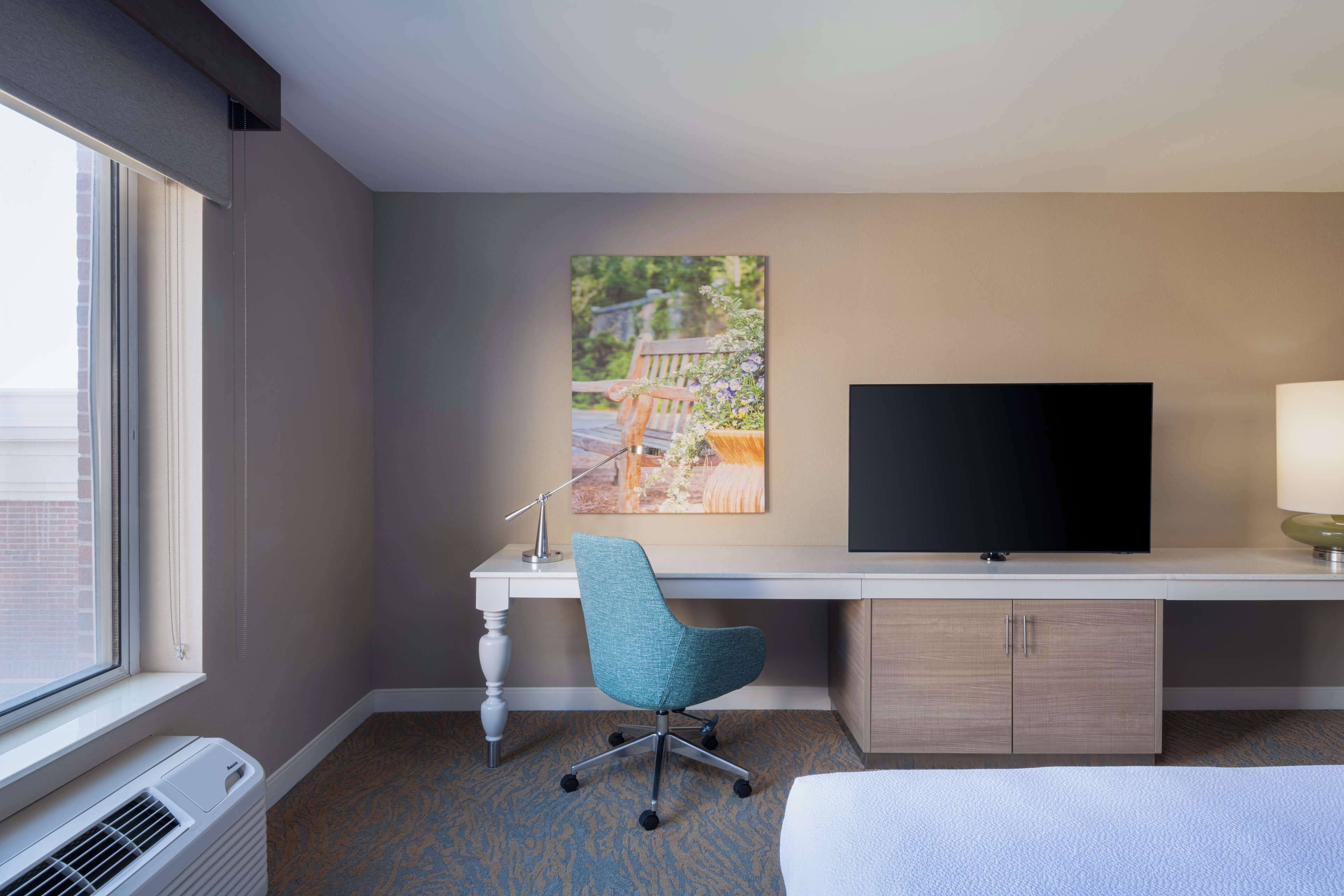 A hotel room with a large window, a desk with a flat-screen TV, a blue chair, and a bed.