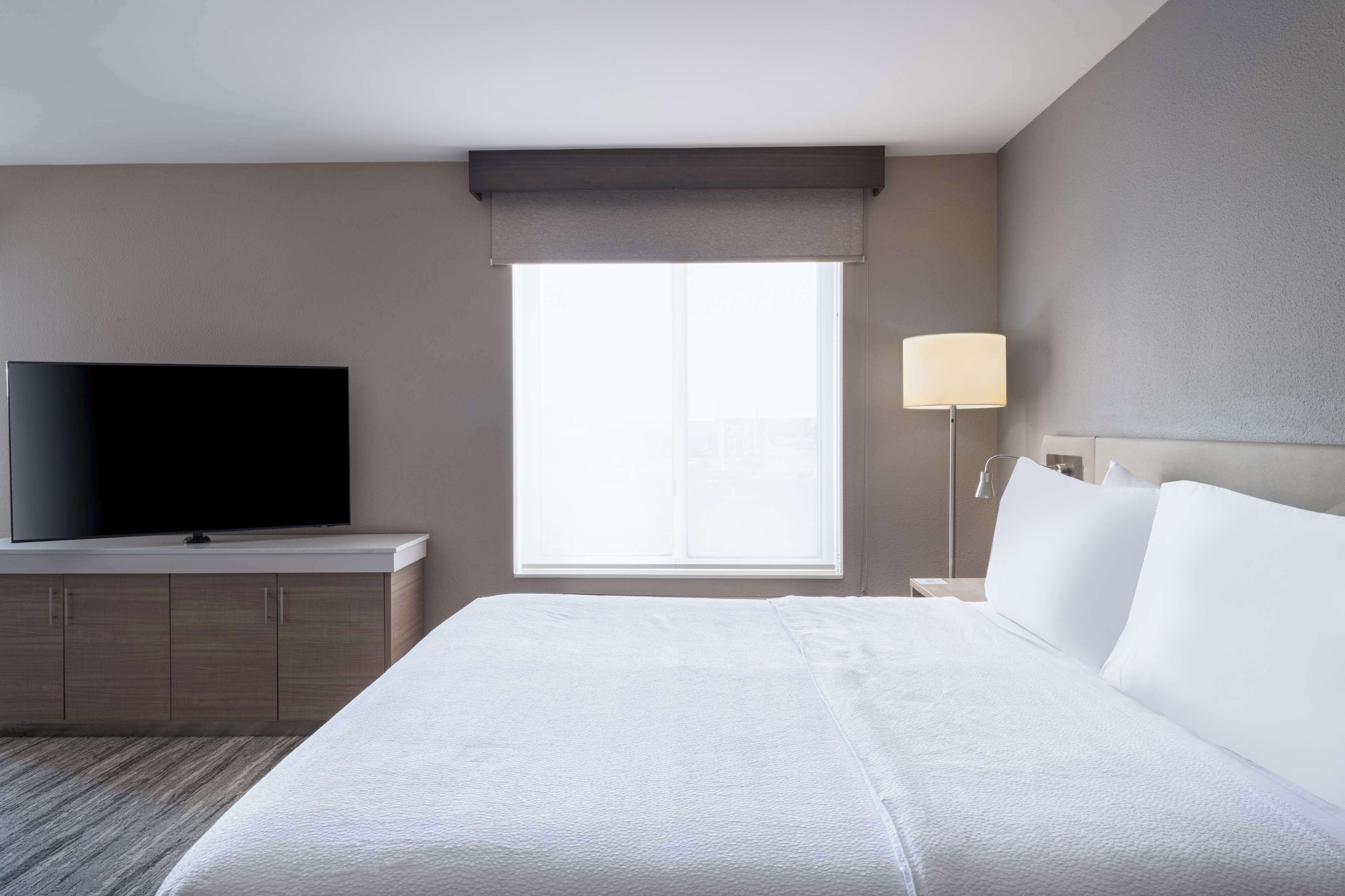 A hotel room with a large bed, a flat-screen TV on a wooden dresser, a window with a view, and a bedside lamp.