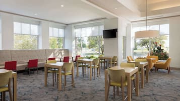Een restaurantgedeelte in een hotel met meerdere tafels en stoelen, grote ramen en een televisie aan de muur.