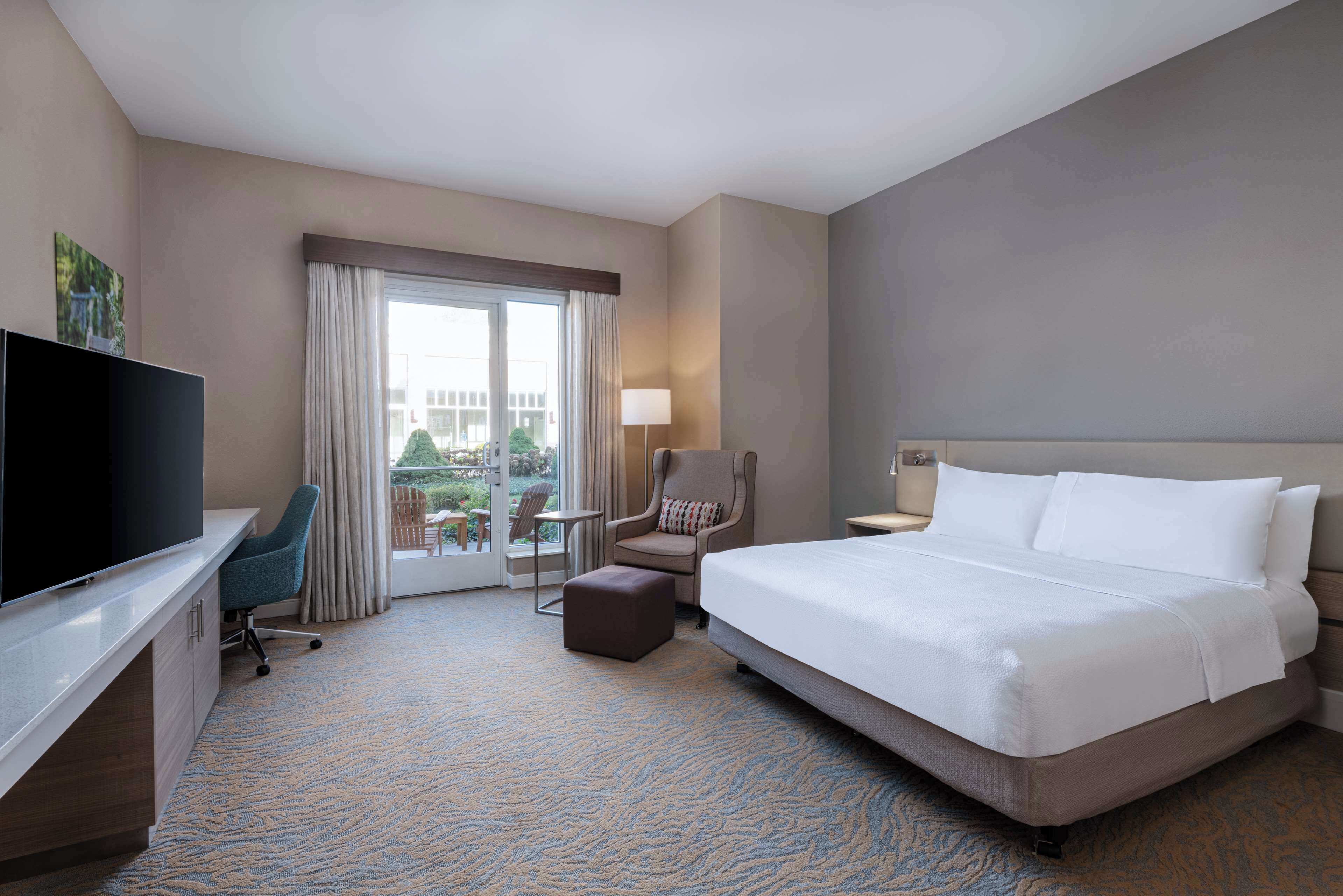 Une chambre d’hôtel avec un grand lit, un bureau avec une télévision, une chaise, une petite table et une fenêtre avec des rideaux.