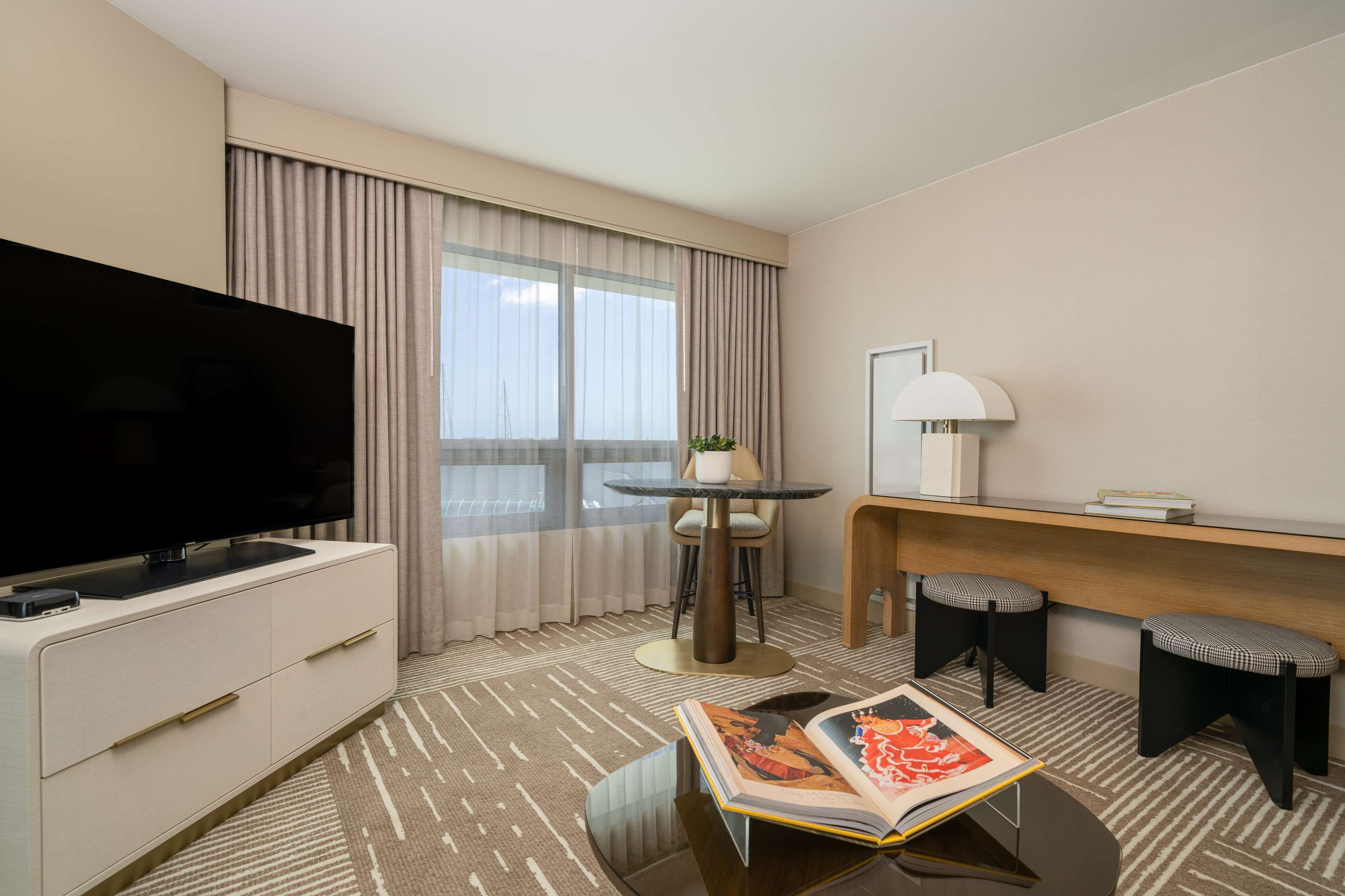 Ein Hotelzimmer mit Flachbildfernseher, einem kleinen runden Tisch mit Lampe, zwei Hocker und einem großen Fenster mit Vorhängen.