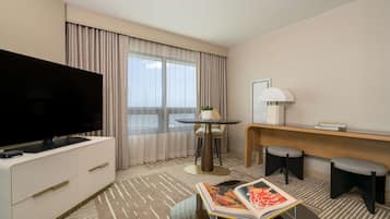 Ein Hotelzimmer mit Flachbildfernseher, einem kleinen runden Tisch mit Lampe, zwei Hocker und einem großen Fenster mit Vorhängen.