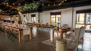 A spacious hall with long tables set for dining, wooden chairs, and hanging plants.