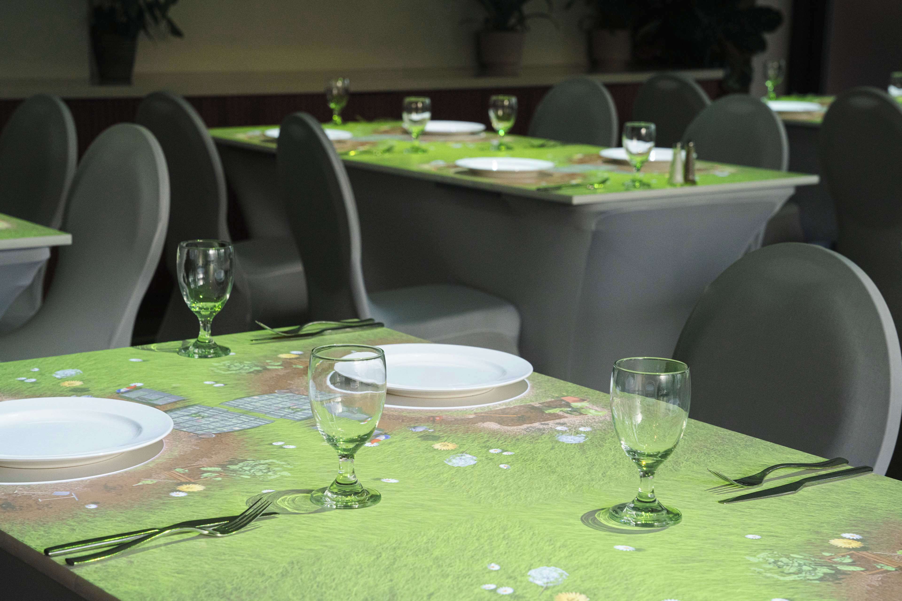 A dining area with tables set for two, each with a green tablecloth, white plates, and clear glassware.