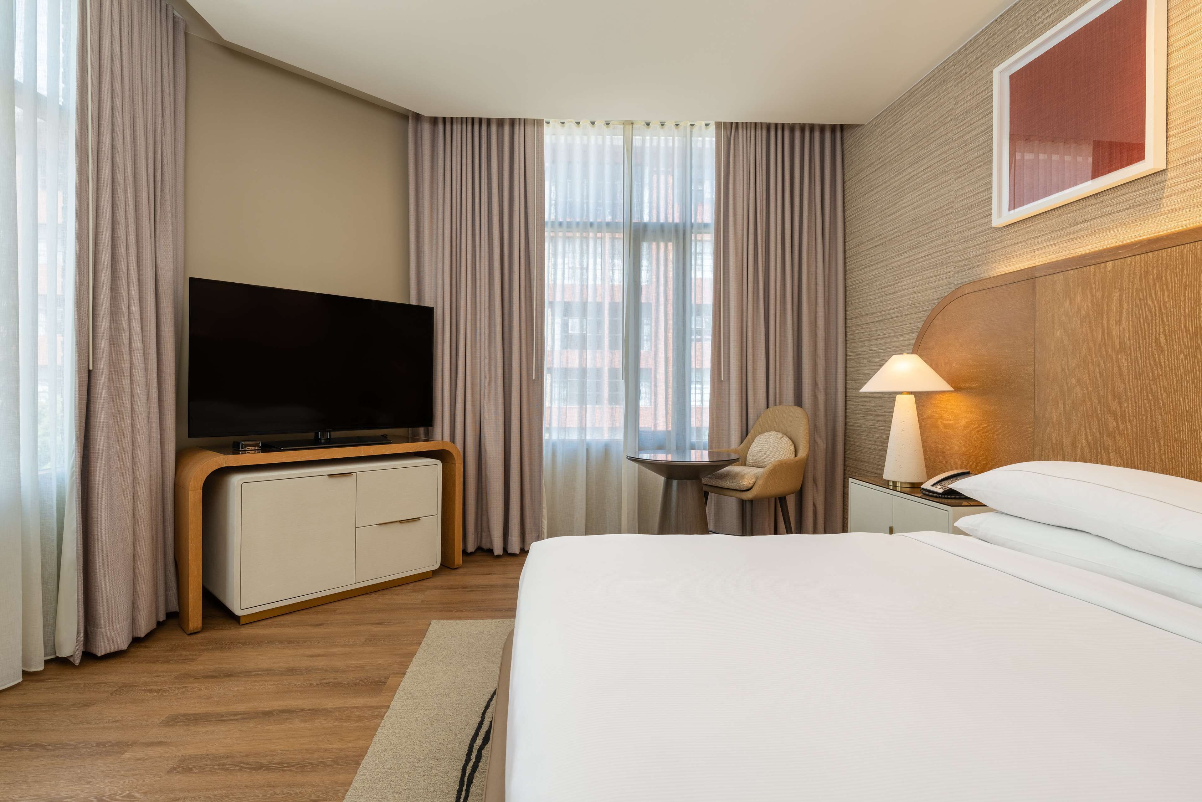 Habitación de hotel con una cama, un televisor sobre un mueble de madera, una mesita de noche con una lámpara y una silla.