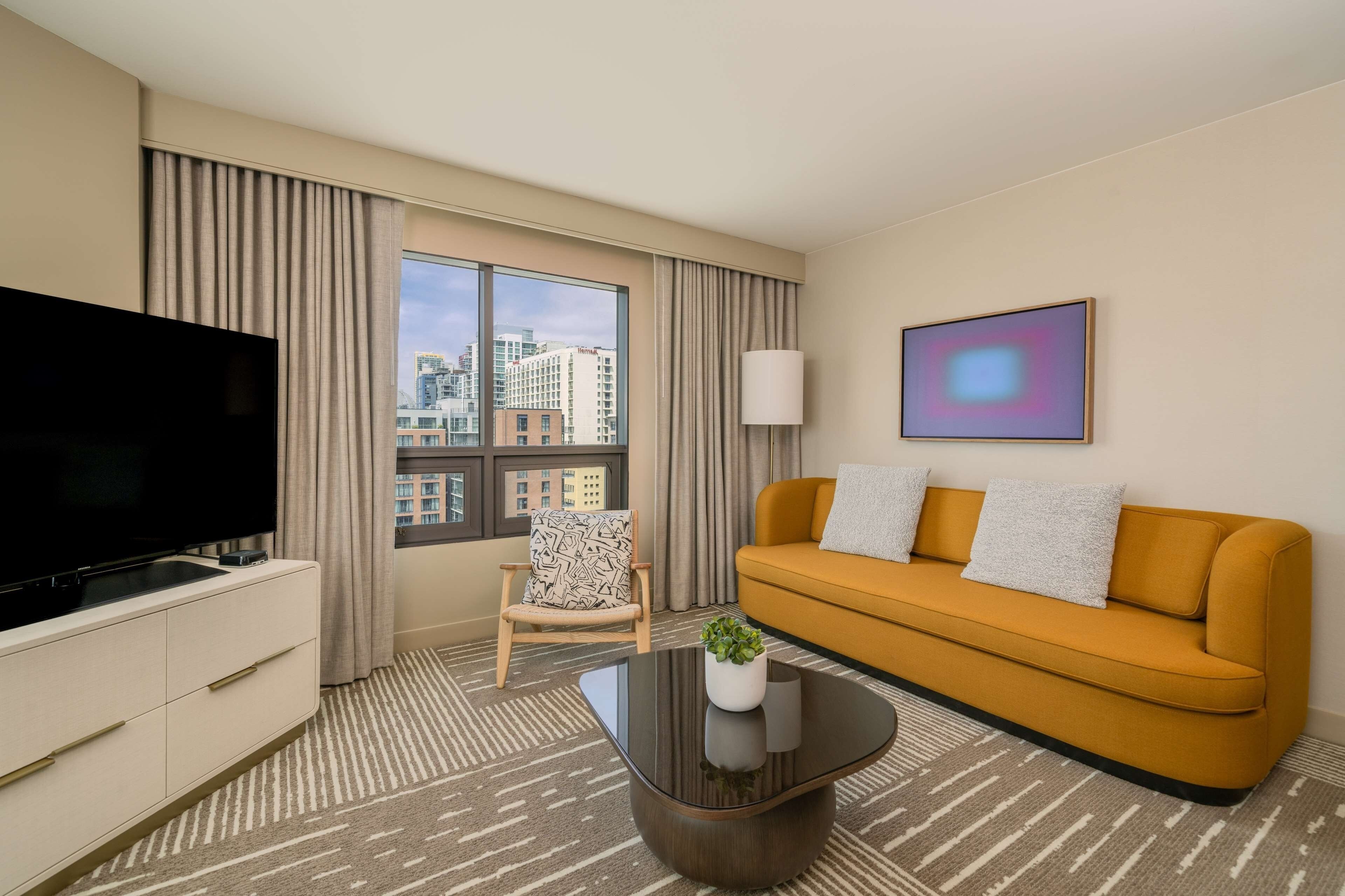 A hotel room with a large TV, a sofa, a chair, and a window with a city view.