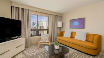 A hotel room with a large TV, a sofa, a chair, and a window with a city view.