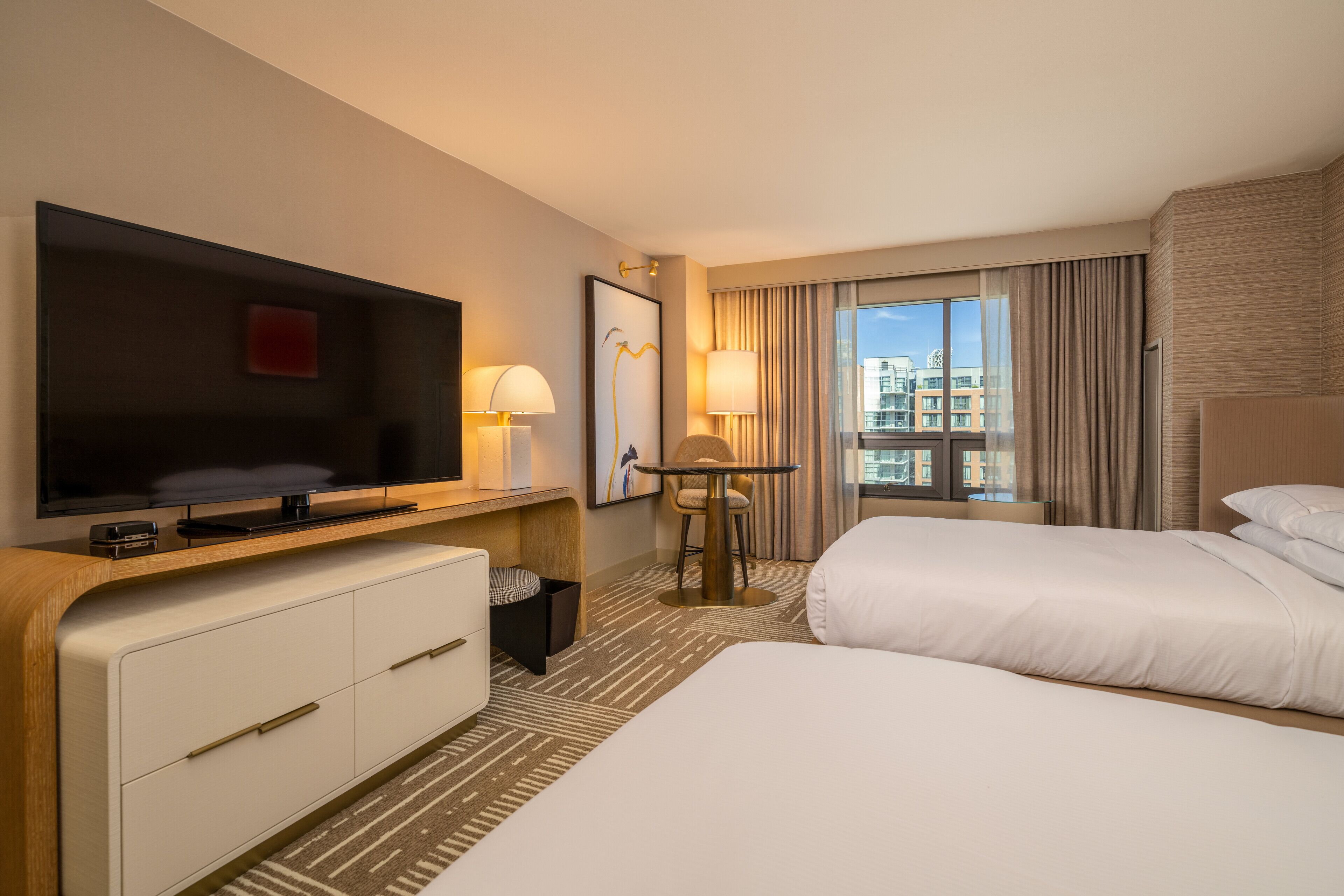 A hotel room with a large flat-screen TV, two beds, a desk with a chair, and a window with curtains.