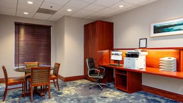 A conference room with a wooden desk, a computer, and a printer. There is a window with blinds, a chair, and a table with chairs.