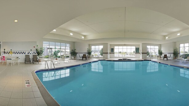 An indoor swimming pool with clear blue water, surrounded by lounge chairs and tables, large windows, and a tiled floor with a 'No Diving' sign. - Hilton Garden Inn Milwaukee Northwest Conference Center (Milwaukee)