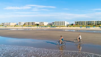 Una playa con dos ciclistas, una orilla arenosa y edificios al fondo.