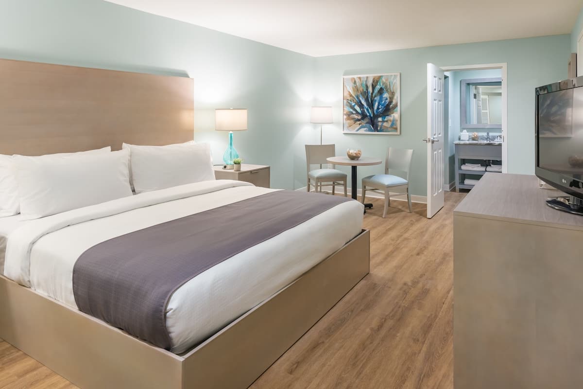 Habitación de hotel con cama, mesitas de noche, escritorio con silla, televisor y baño a la vista a través de una puerta abierta.