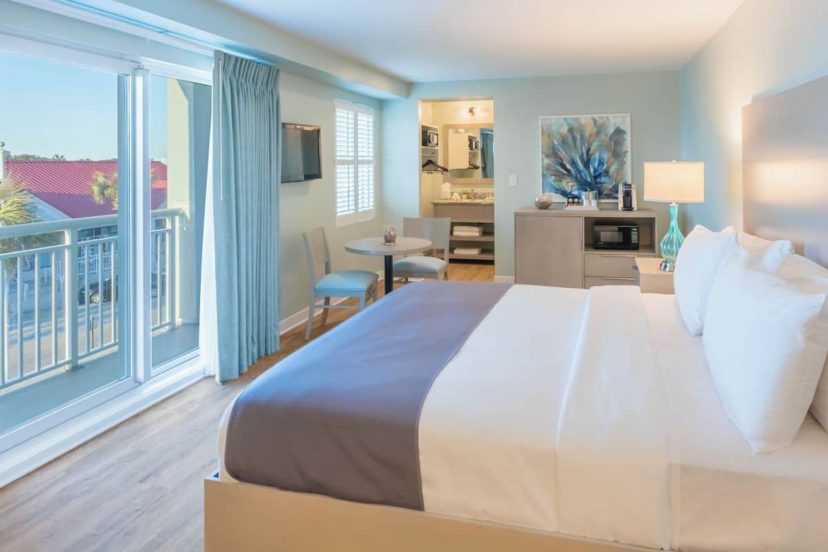 Habitación de hotel con una cama grande, un escritorio, una silla, una pequeña cocina y un balcón con vistas a edificios.