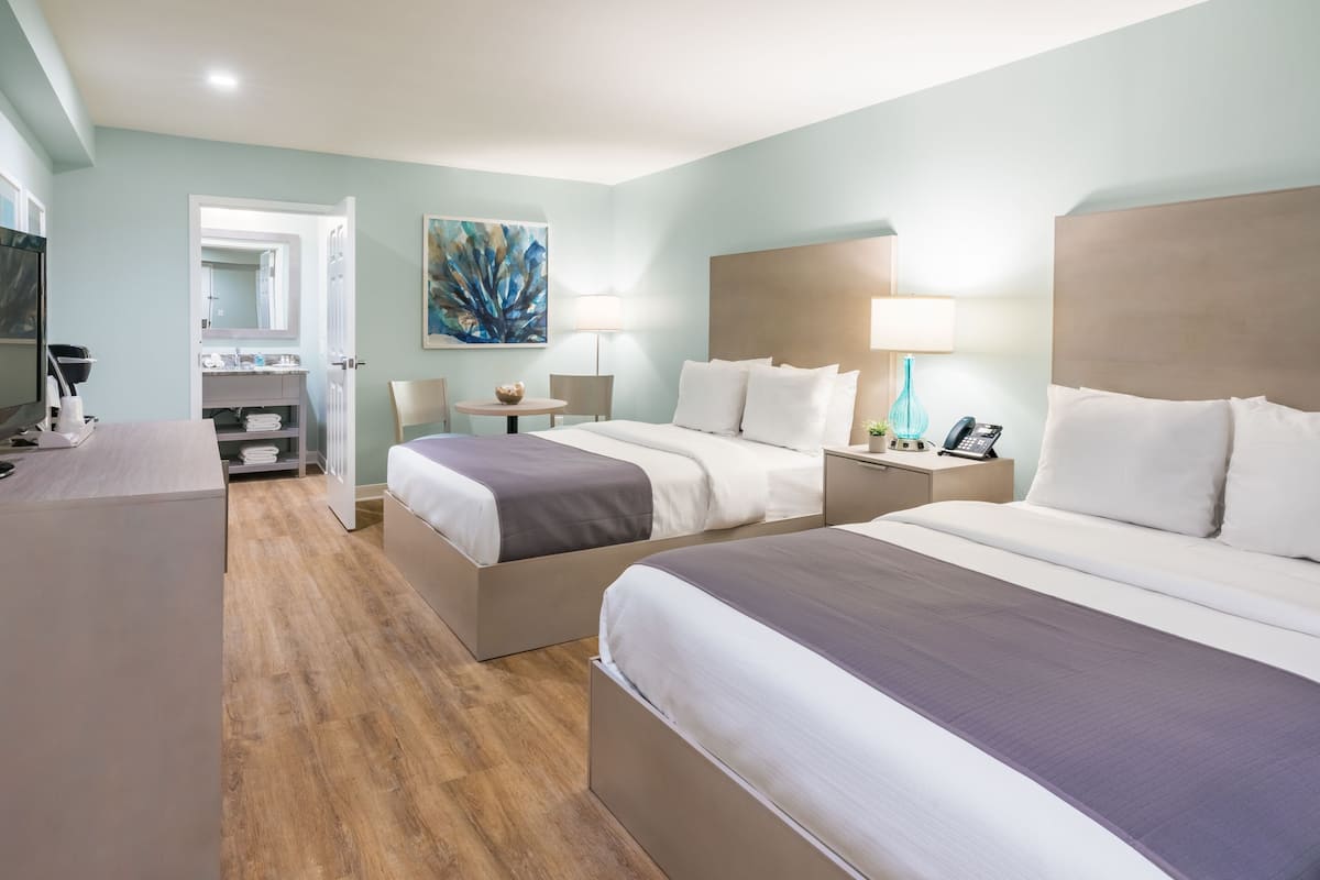 Habitación de hotel con dos camas, un escritorio, una silla, un televisor y un baño visible a través de una puerta abierta.