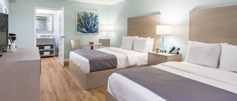 Habitación de hotel con dos camas, un escritorio, una silla, un televisor y un baño visible a través de una puerta abierta.