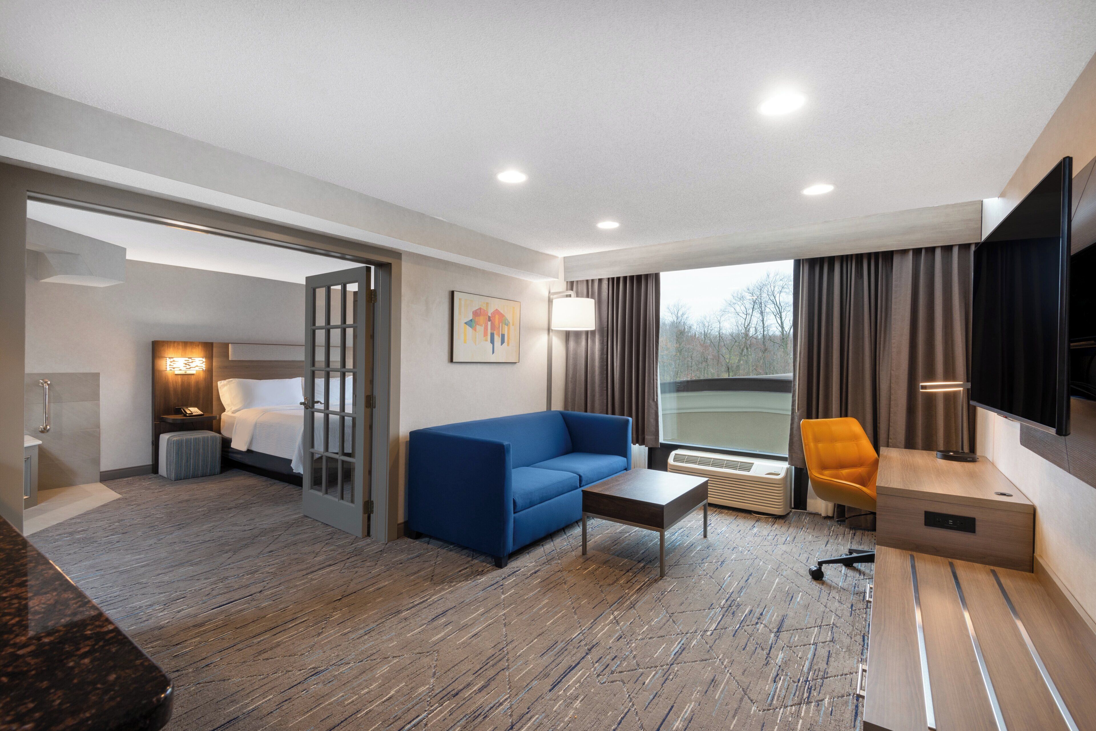 Ein Hotelzimmer mit einem Bett, einem blauen Sofa, einem Couchtisch, einem Fernseher, einem Sessel und einem fenster mit Vorhängen.