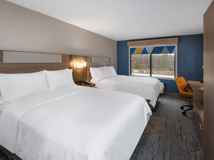 A hotel room with two beds, a desk, a chair, and a window with a view of trees.