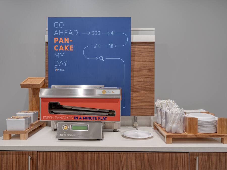 A breakfast station with a flat-screen TV displaying a breakfast menu, a pancake maker, and a coffee machine.