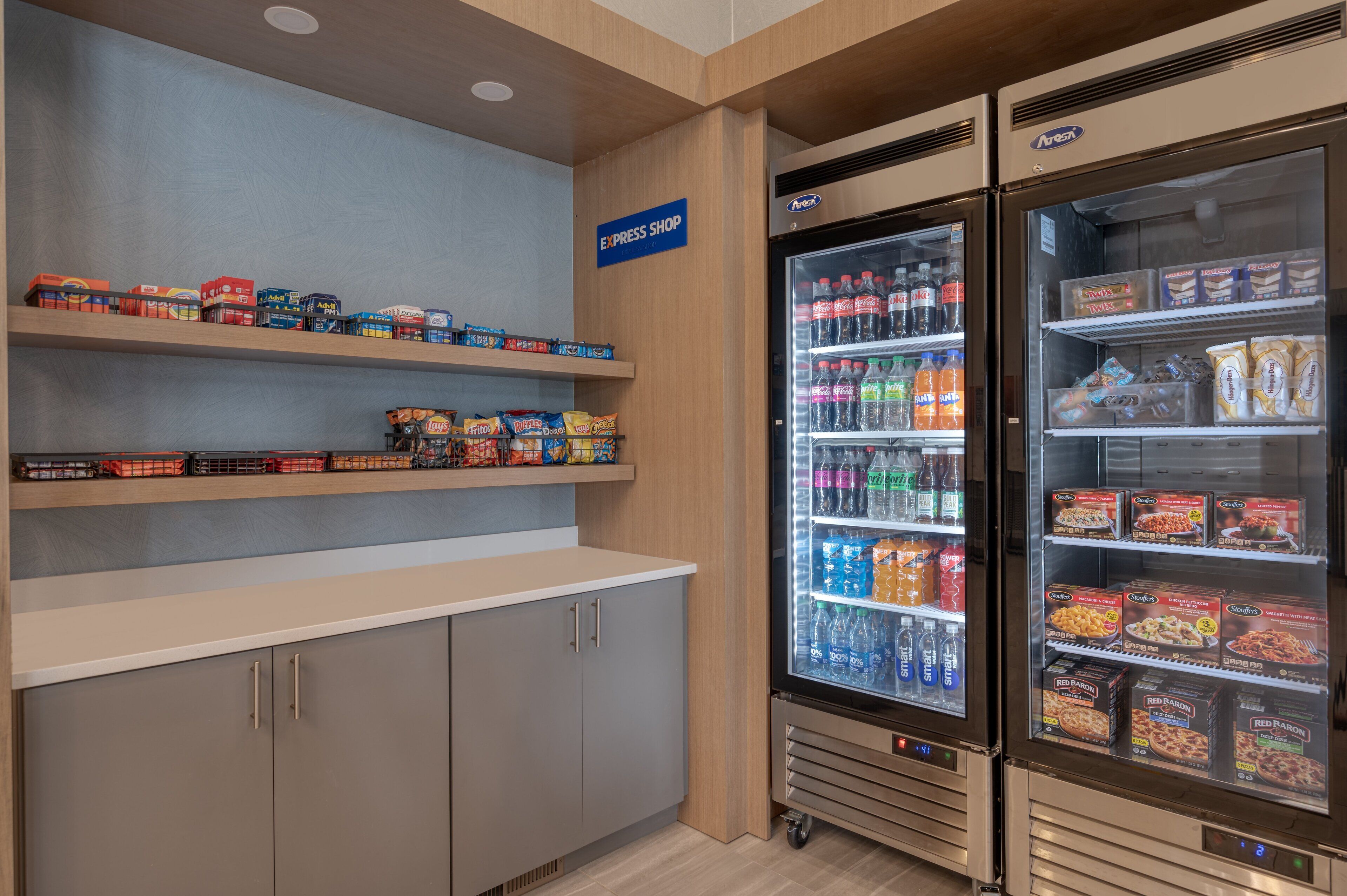 A self-service snack area with shelves stocked with various snacks and a refrigerated display case with beverages and packaged foods.