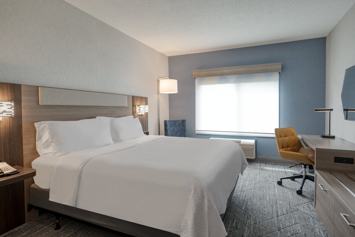 A hotel room with a large bed, a desk, a chair, and a window with blinds.