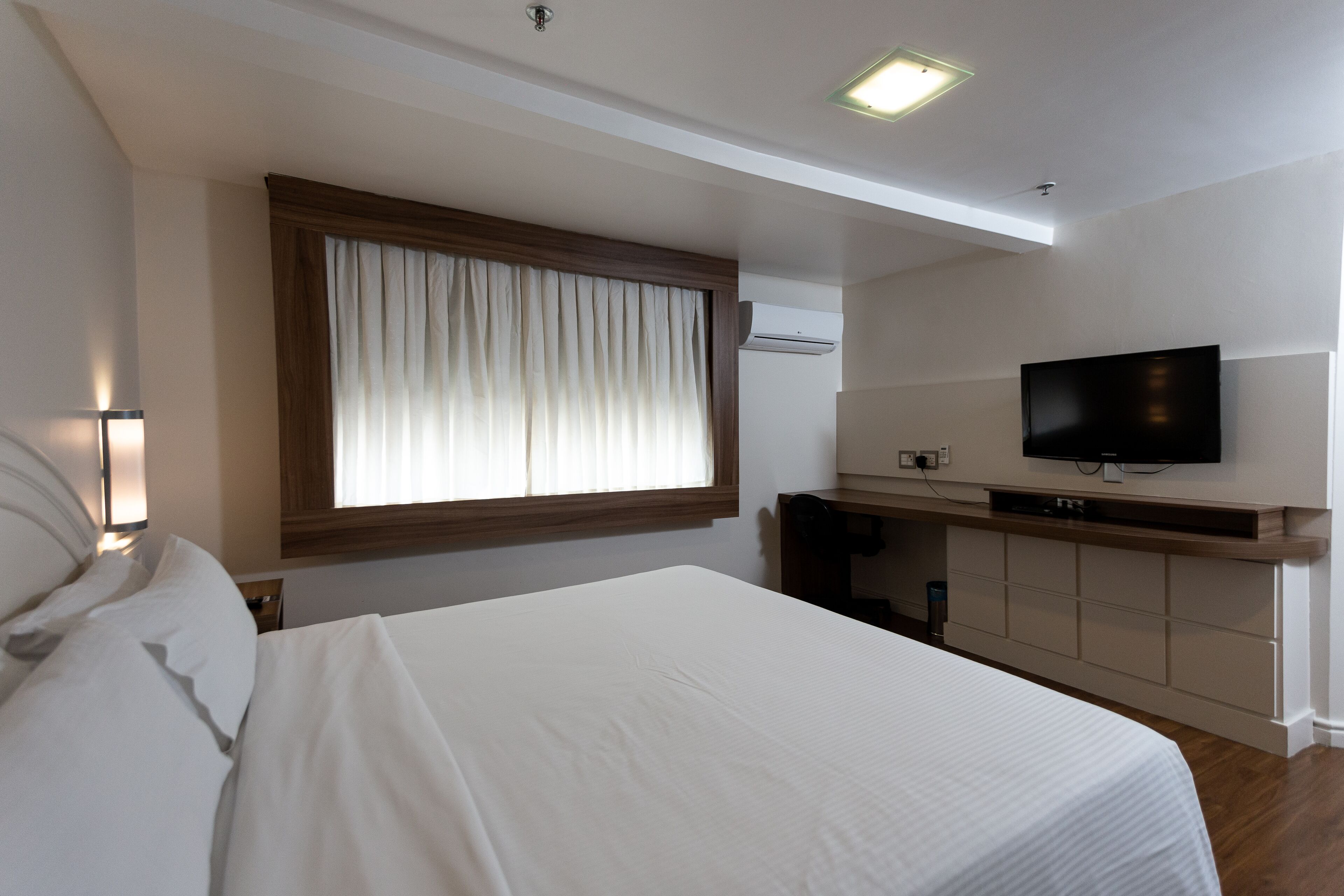 Quarto de hotel com uma cama grande, uma escrivaninha com televisão de tela plana e uma janela com cortinas.