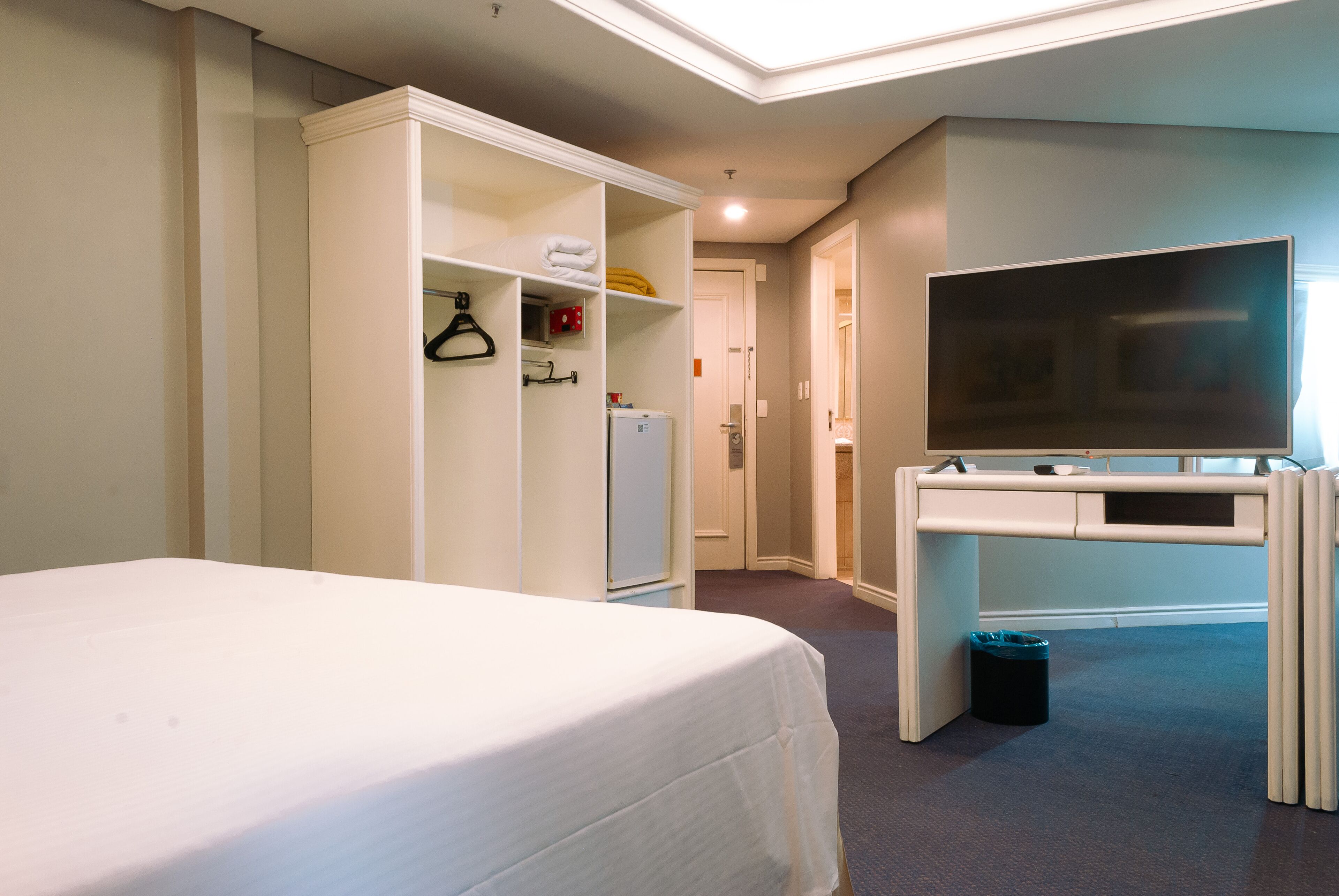 A hotel room with a bed, a wardrobe, a desk with a television, and a trash bin.