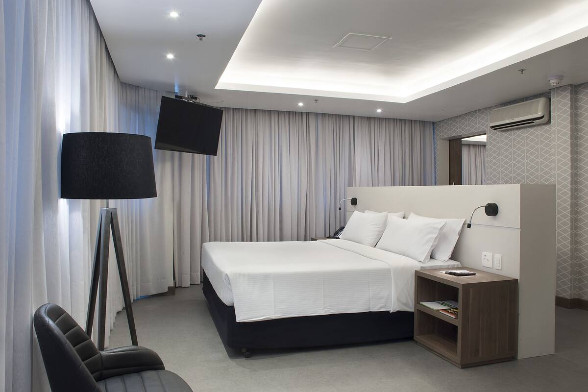 A modern hotel room with a large bed, a desk with a lamp, and a flat-screen TV mounted on the wall.