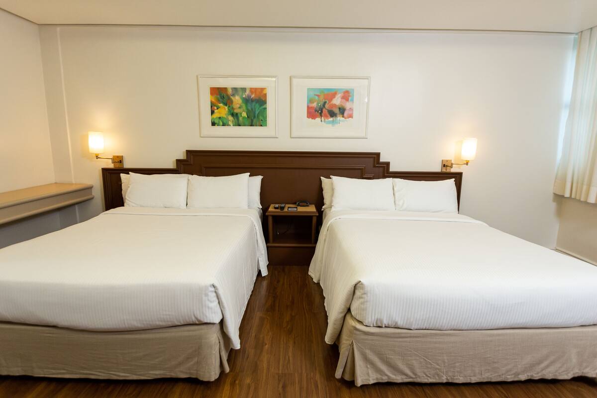 A hotel room with two single beds, a wooden headboard, bedside tables with lamps, and two framed artworks on the wall.