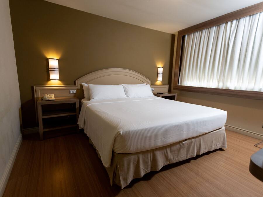 Habitación de hotel con una cama grande, dos mesitas de noche, un buró, una ventana con cortinas y piso de madera.