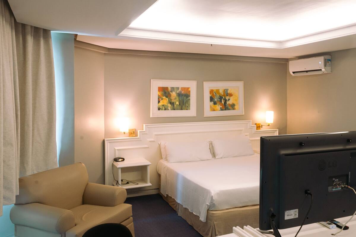 A hotel room with a bed, a TV, a chair, a desk, and two paintings on the wall.