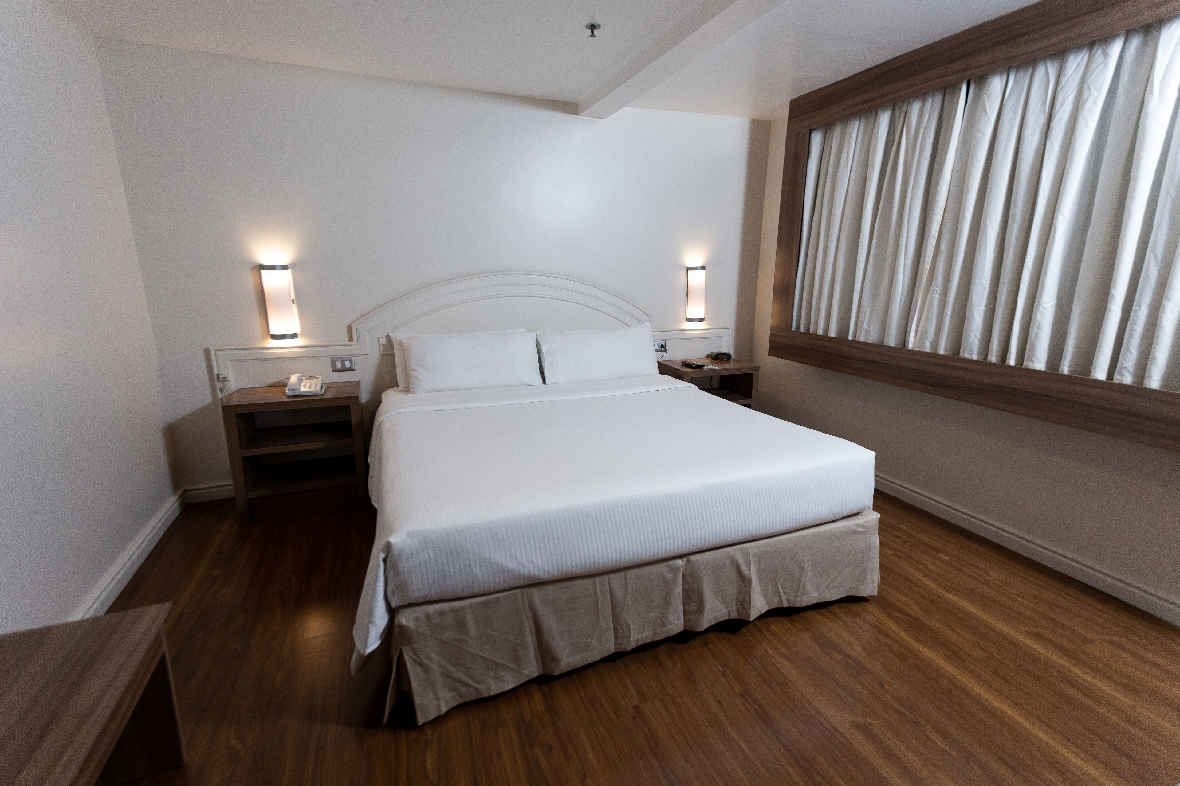 A hotel room with a large bed, wooden flooring, two bedside tables with lamps, and a window with curtains.