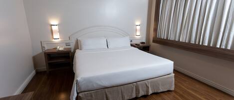 A hotel room with a large bed, wooden flooring, two bedside tables with lamps, and a window with curtains.