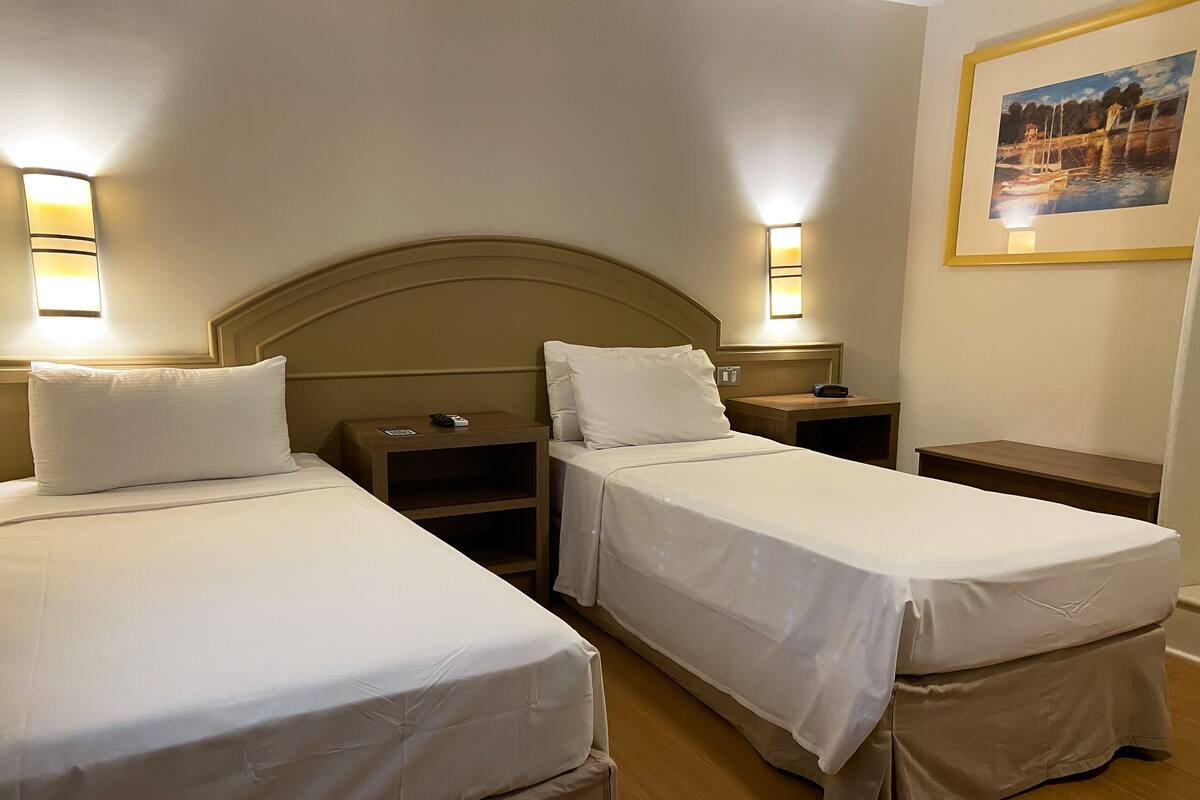A hotel room with two single beds, each with a headboard, bedside tables, and lamps.