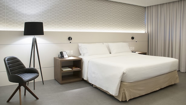 A modern hotel room with a neatly made bed, a chair, a bedside table with a lamp, and a geometric-patterned wall. - Master Porto Alegre Hotel - Av Carlos Gomes, Proximo Consulado Americano (Porto Alegre)