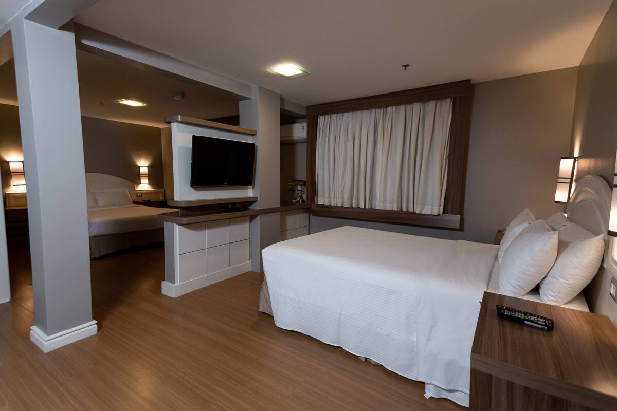 A hotel room with a bed, a TV, a desk, and a window with curtains.