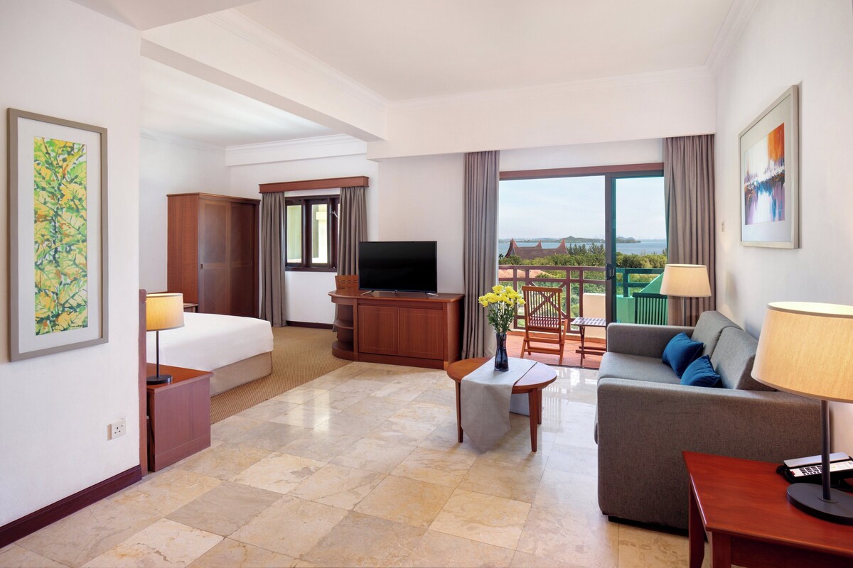 A hotel room with a bed, a sofa, a TV, a desk with a lamp, and a view of the outdoors.