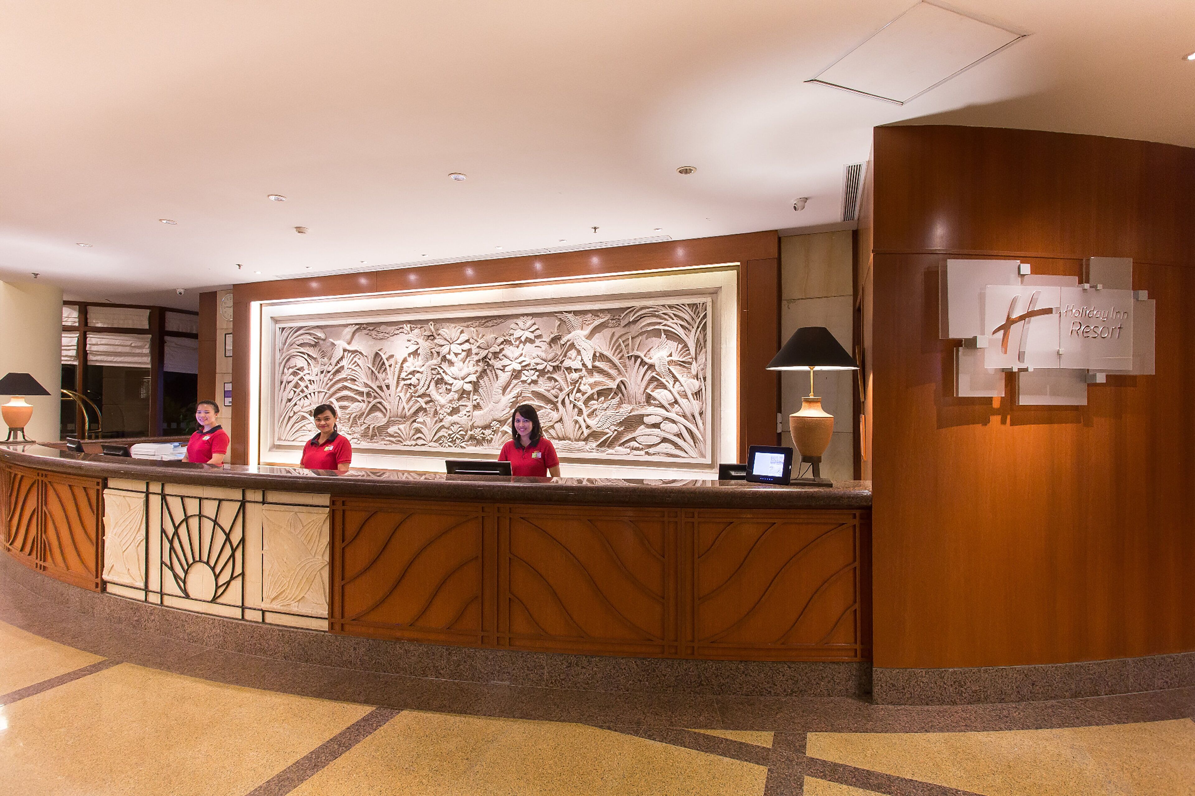A reception desk with a large floral carving, two receptionists, and a sign for Holiday Inn Resort.