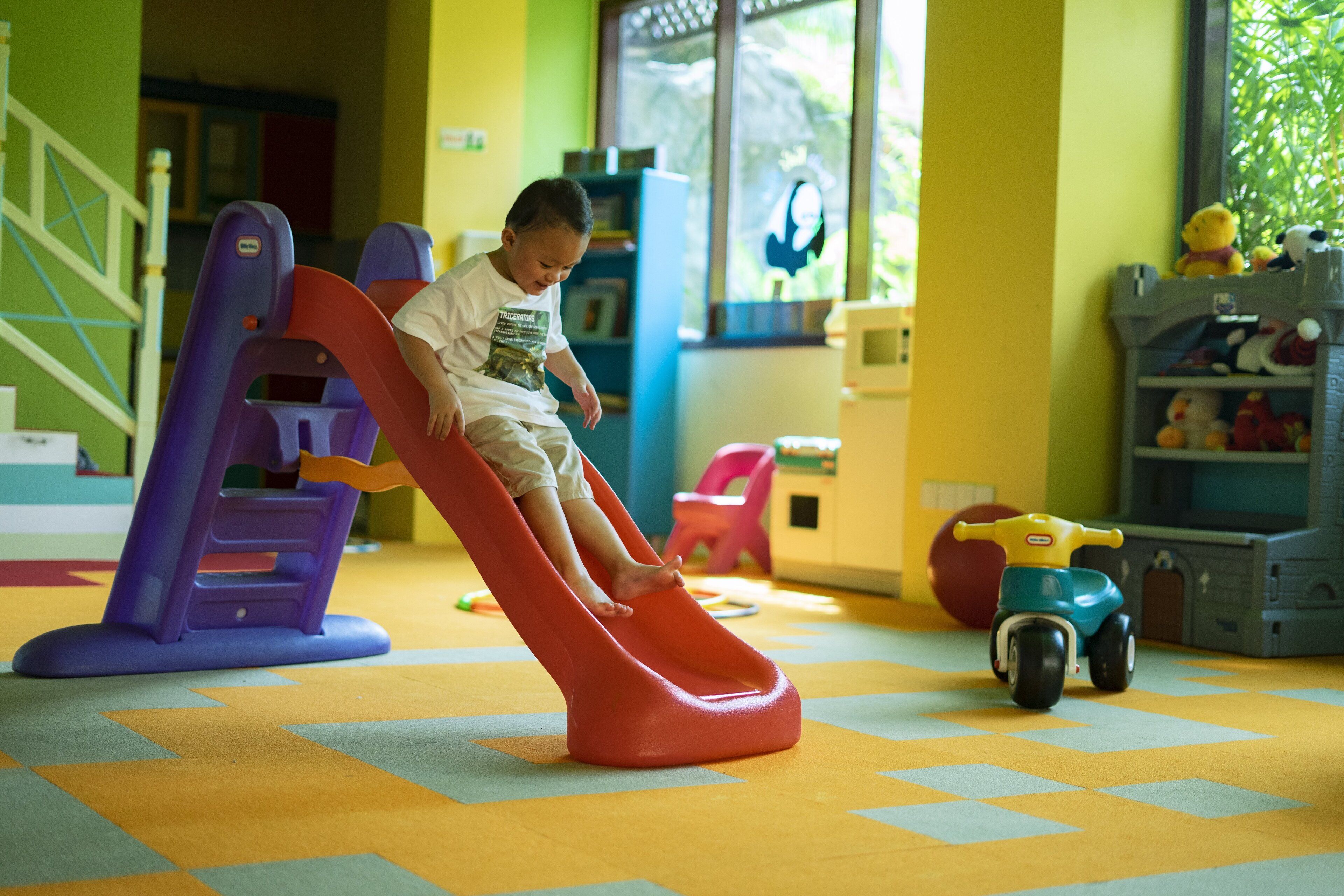 Un niño jugando en un tobogán dentro de una habitación de colores vivos, con un coche de juguete y estanterías al fondo.