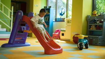 Un niño jugando en un tobogán dentro de una habitación de colores vivos, con un coche de juguete y estanterías al fondo.