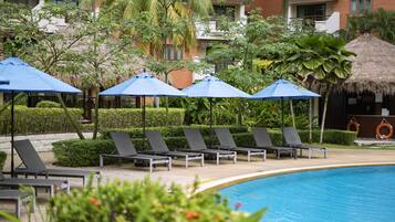 A poolside area with lounge chairs and umbrellas, surrounded by greenery and a building with a thatched roof.