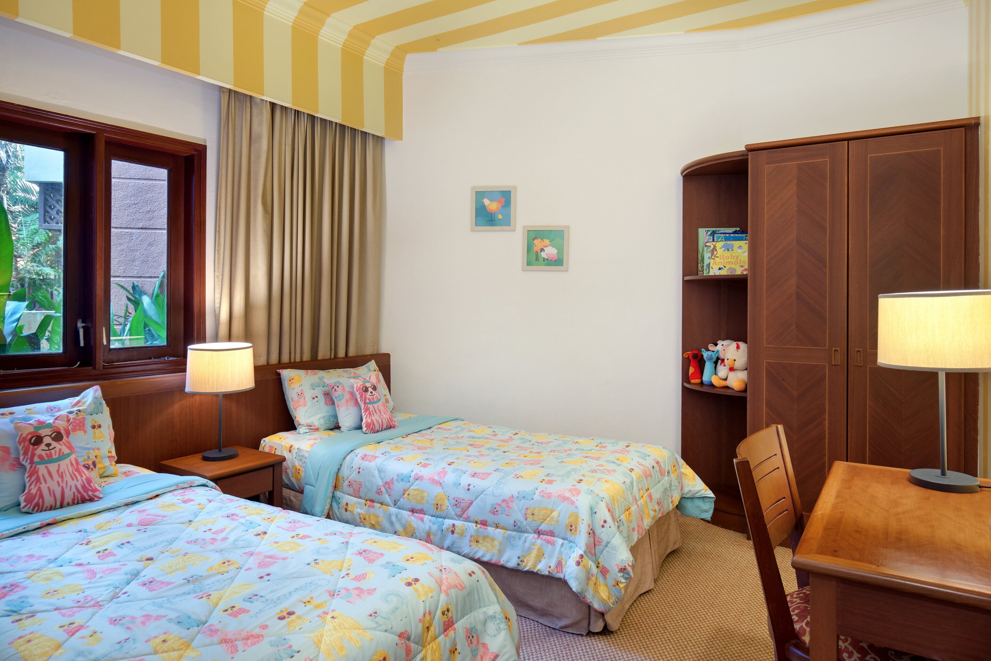A bedroom with two beds, a wooden wardrobe, a desk with a lamp, and a window with curtains.
