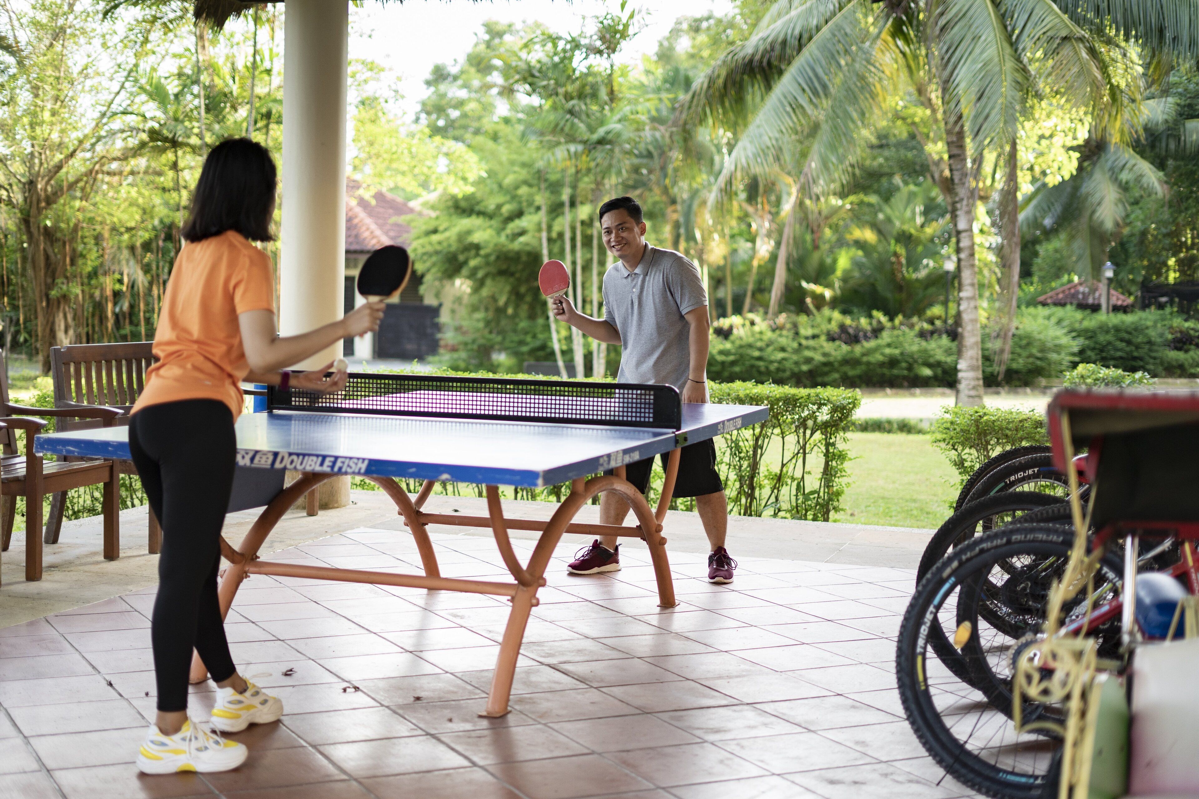 Mesa de ping pong instalada al aire libre donde una pareja juega, rodeada de bicicletas y follaje.