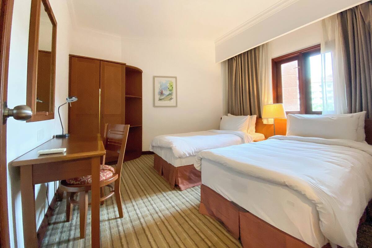 Habitación de hotel con dos camas, un escritorio, una silla, una lámpara y un cuadro en la pared.