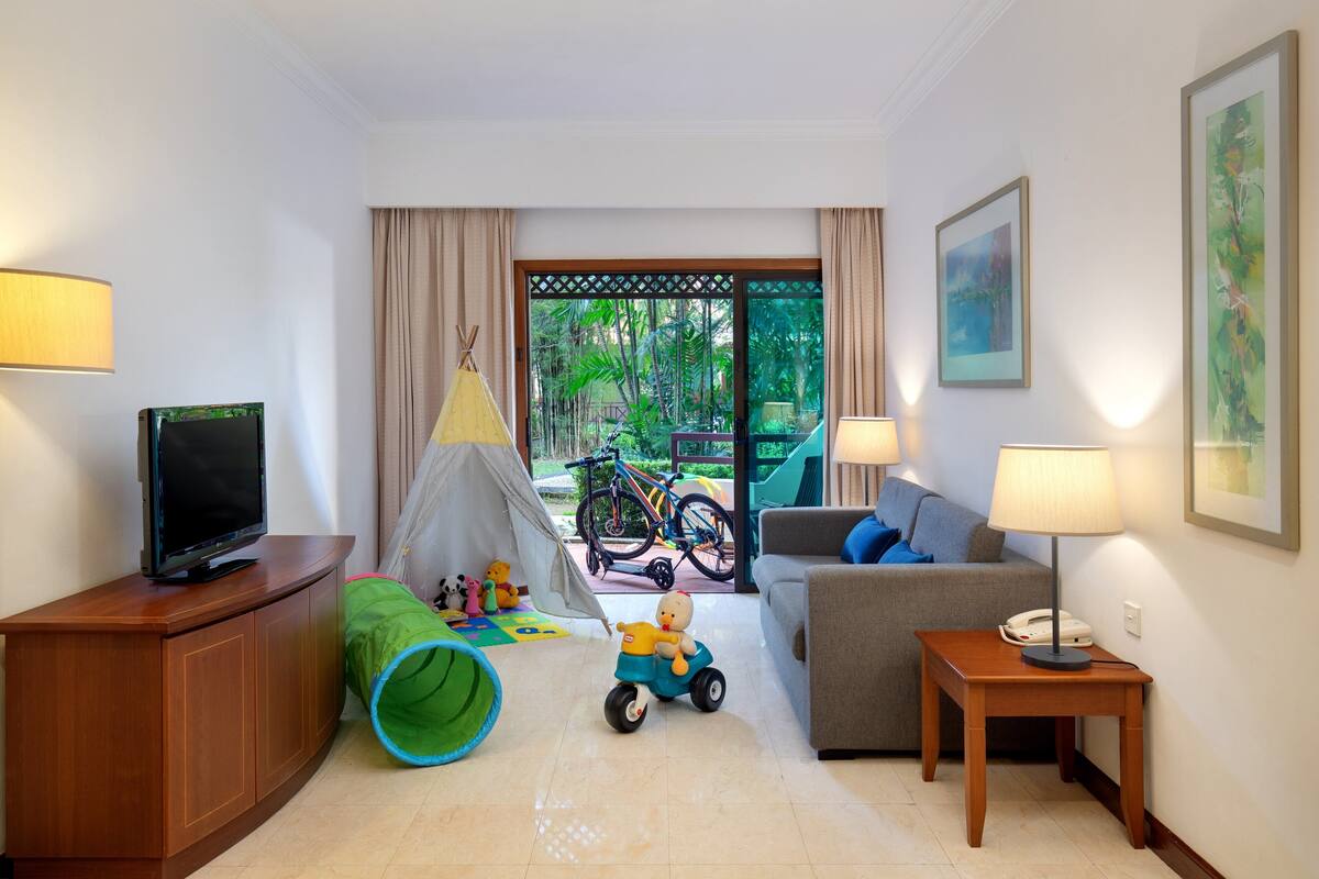 A hotel room with a television, a sofa, a small table with a lamp, a green tunnel play area, a tent, and bicycles outside the window.