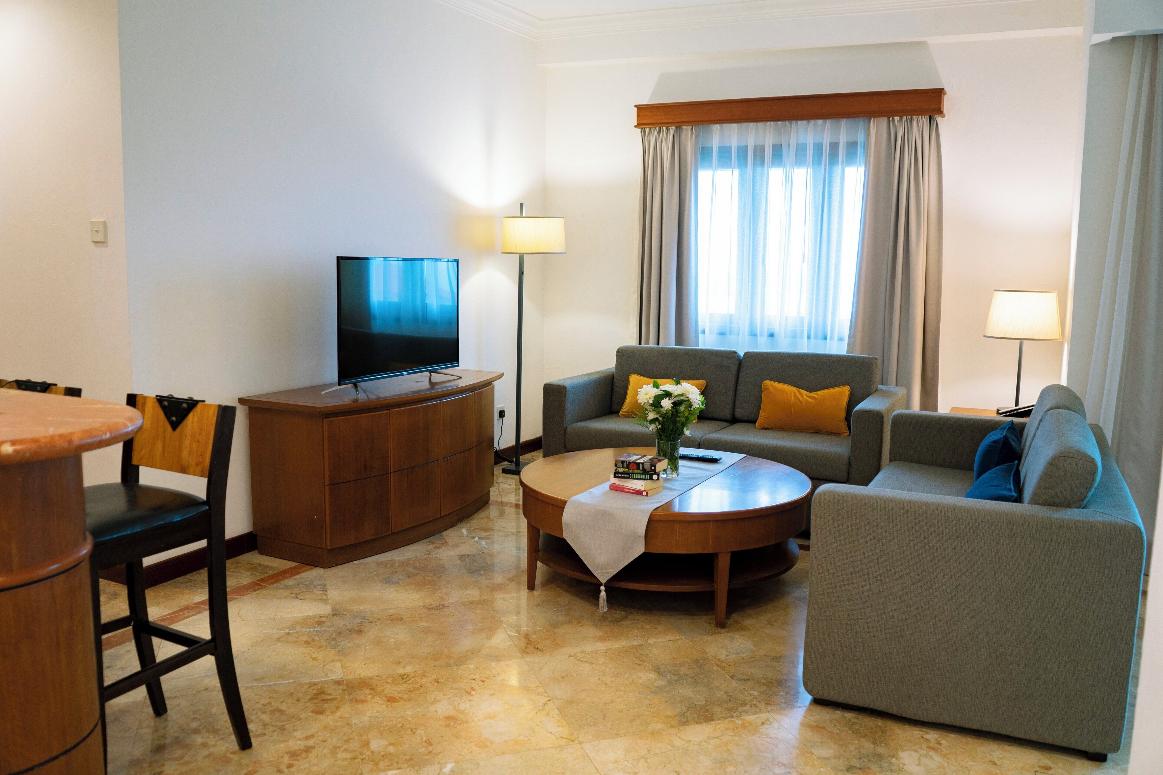 A modern hotel room with a flat-screen TV, a sofa, a coffee table with a vase and books, and a dining area with a wooden table and chairs.