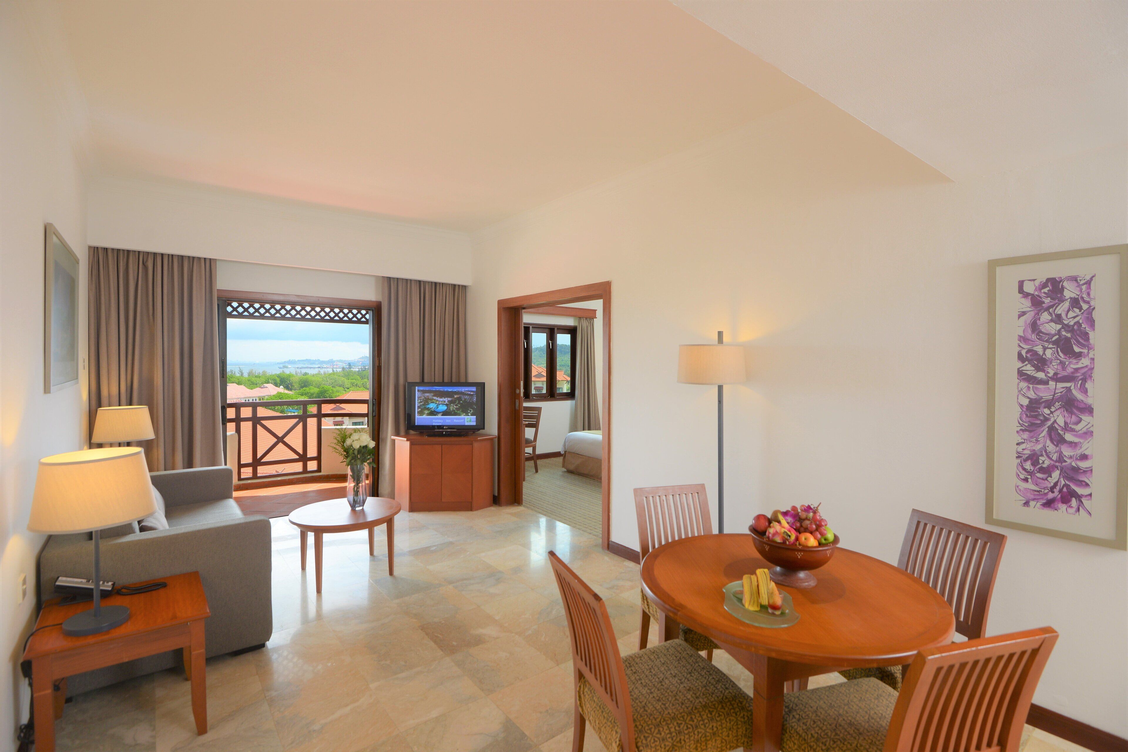 A hotel room with a sofa, TV, dining table, chairs, and a view of the outdoors.