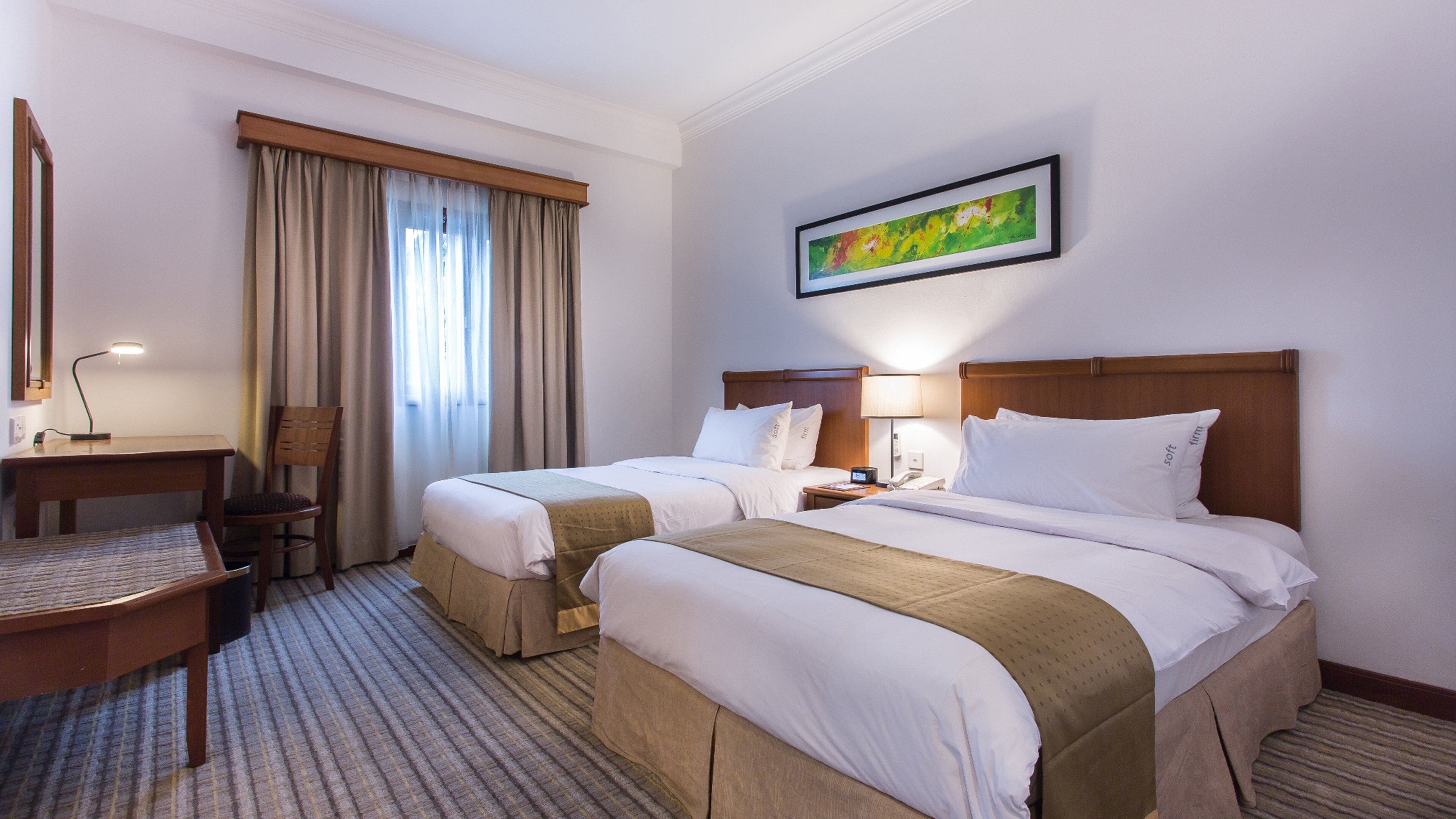 A hotel room with two beds, a desk, a chair, a window with curtains, a painting on the wall, and a carpeted floor.